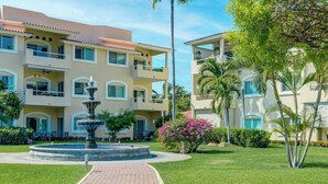 Un hotel de varios pisos con una fuente central, rodeado de céspedes cuidados y palmeras.