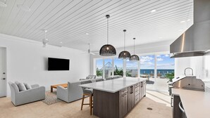 A modern kitchen with a large island, a dining area with a sofa and chairs, and a view of the ocean through large windows. - 'Domus Pacis' | Two Pools, Beach Front,  Next to Seaside 30A (Seagrove Beach)