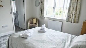 Una cama bien hecha con una colcha estampada y dos toallas dobladas. Una silla con cojín está ubicada cerca de una ventana con cortinas.