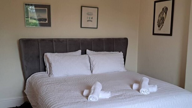 A neatly made bed with white linens and towels, a grey upholstered headboard, and framed artwork on the walls. - Stunning 3BD Victorian House in Bramhall Village (Bramhall)