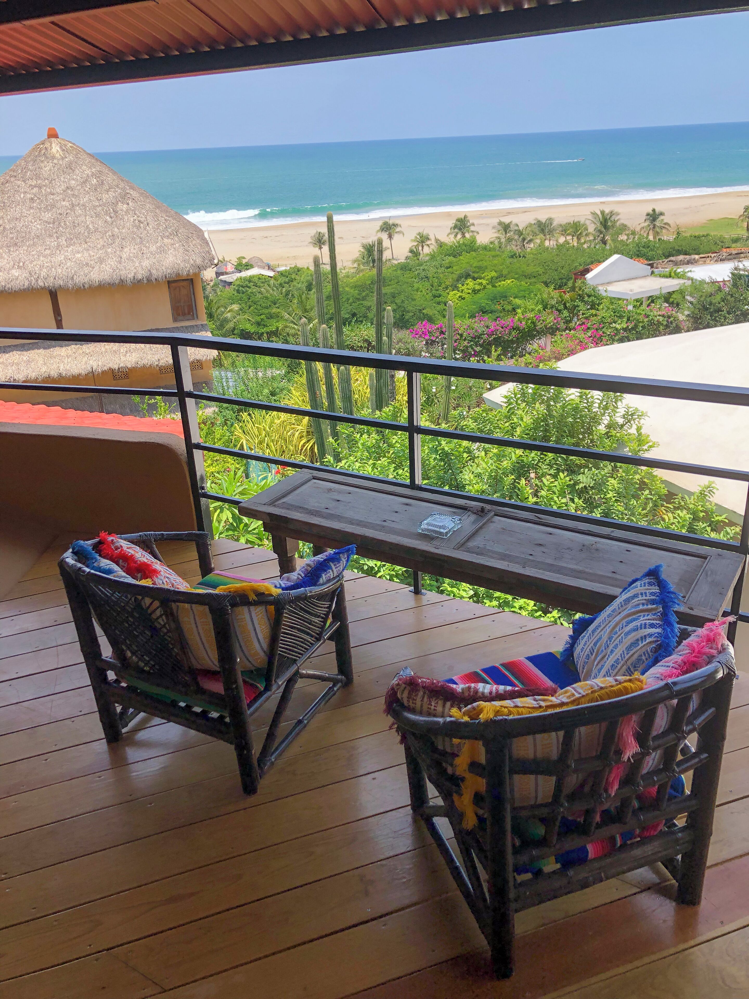 Une terrasse en bois avec deux fauteuils en osier et une table, donnant sur une plage où se trouve une structure à toit de chaume et des cactus.