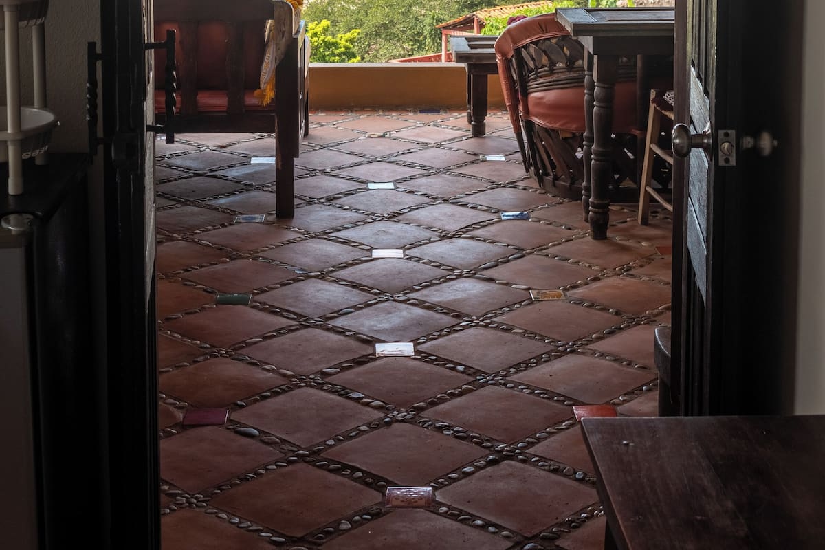 Una porta aperta che dà su un patio con pavimento piastrellato e mobili in legno, con vista su una spiaggia con palme e l'oceano.