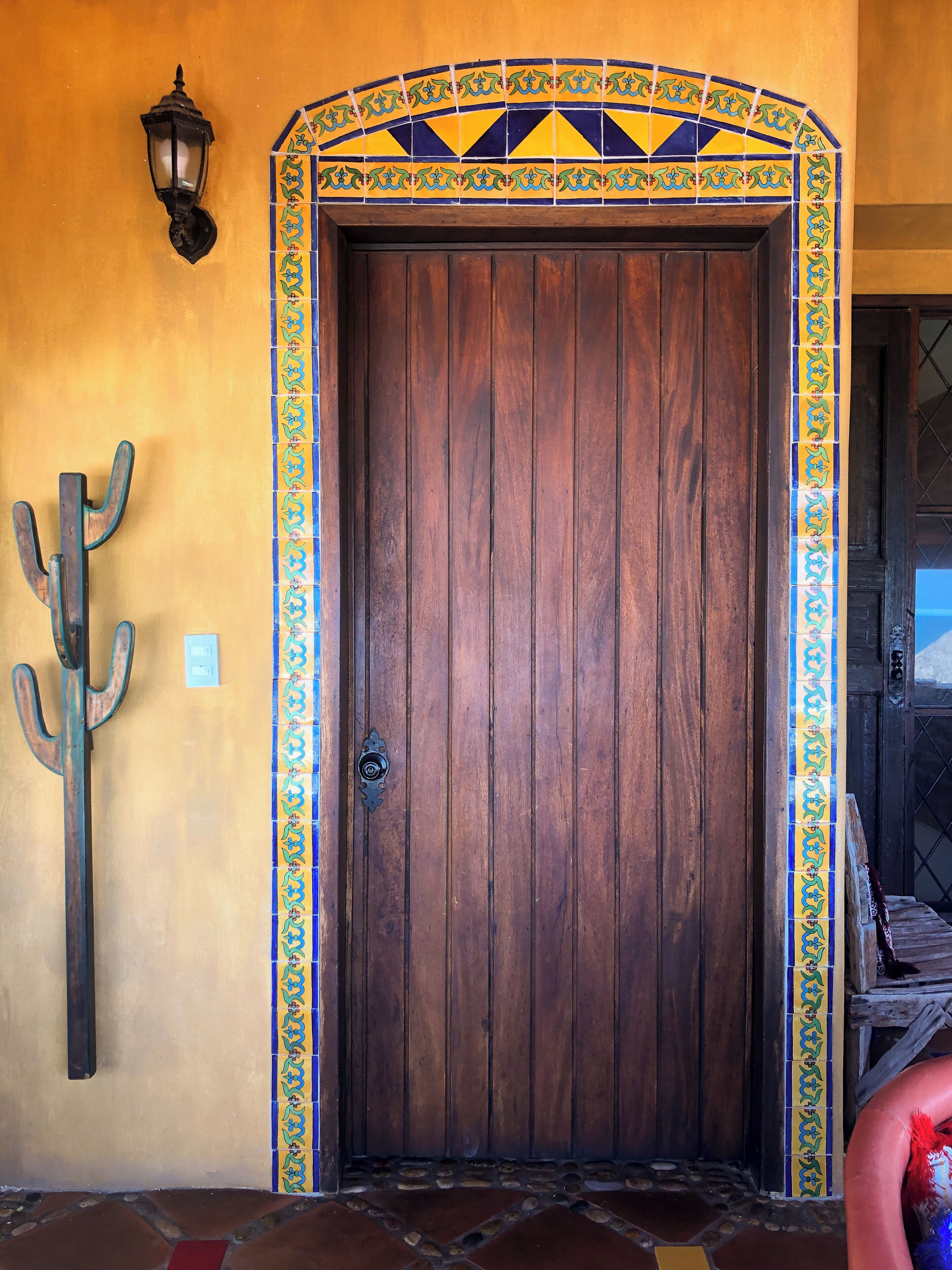 A wooden door with a decorative tiled border, a metal cacti decoration, and a wall-mounted lantern.