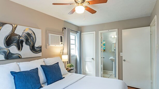 A bedroom with a bed, a ceiling fan, a wall-mounted air conditioner, and a door leading to a bathroom. - The Palm Flats 16! Zoo! Museums! Medical Center! (Houston)