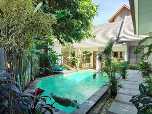 A swimming pool surrounded by lush tropical plants, with a wooden deck and a building in the background. - Manuver Surf and Stay Uluwatu Bali (Jimbaran)