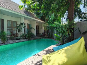 A swimming pool with a wooden deck, surrounded by lush greenery and a house with large windows and a tiled roof. - Manuver Surf and Stay Uluwatu Bali (Jimbaran)