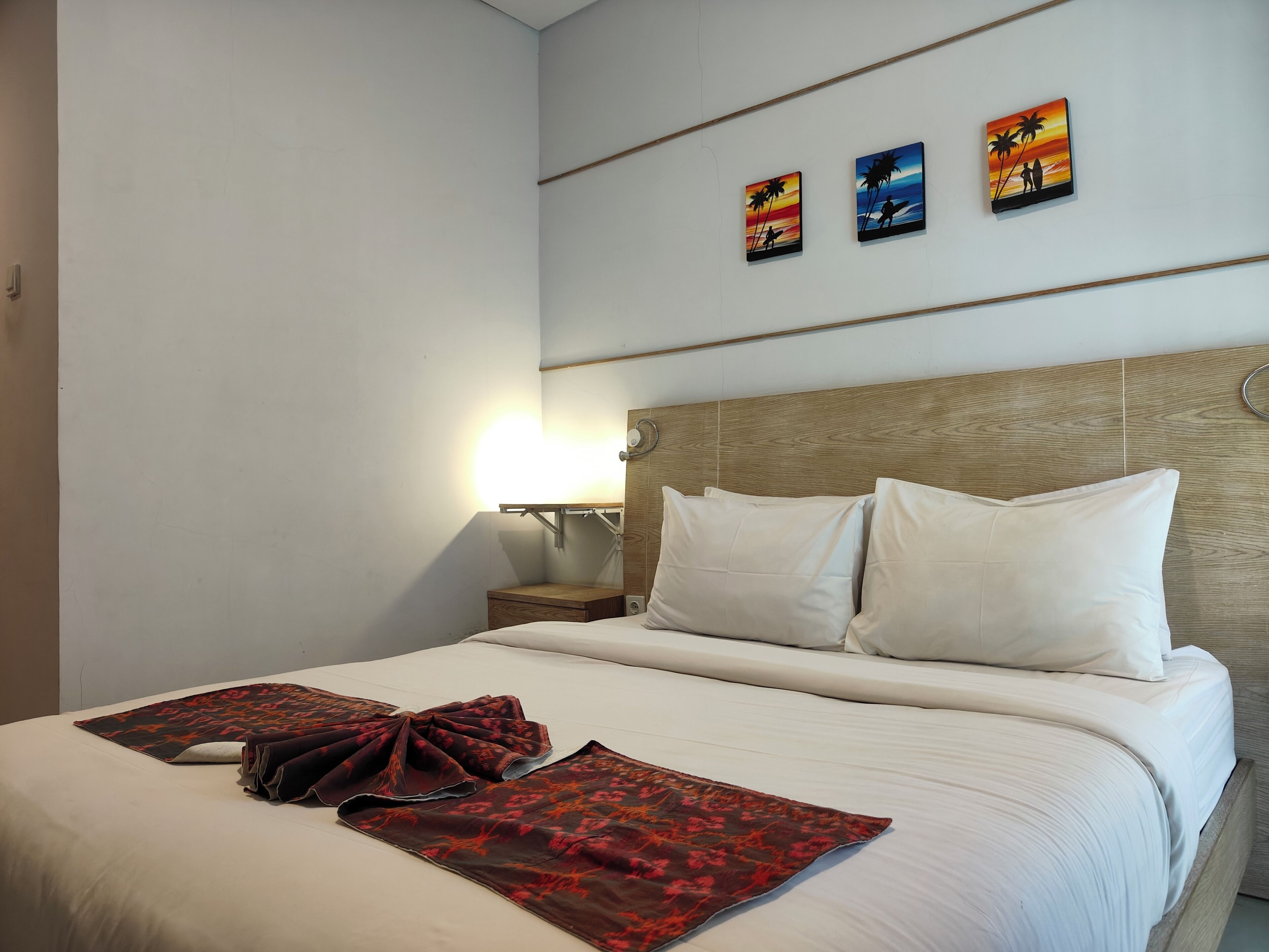 Habitación de hotel con una cama, dos almohadas, una mesita de noche con lámpara y tres cuadros en la pared.