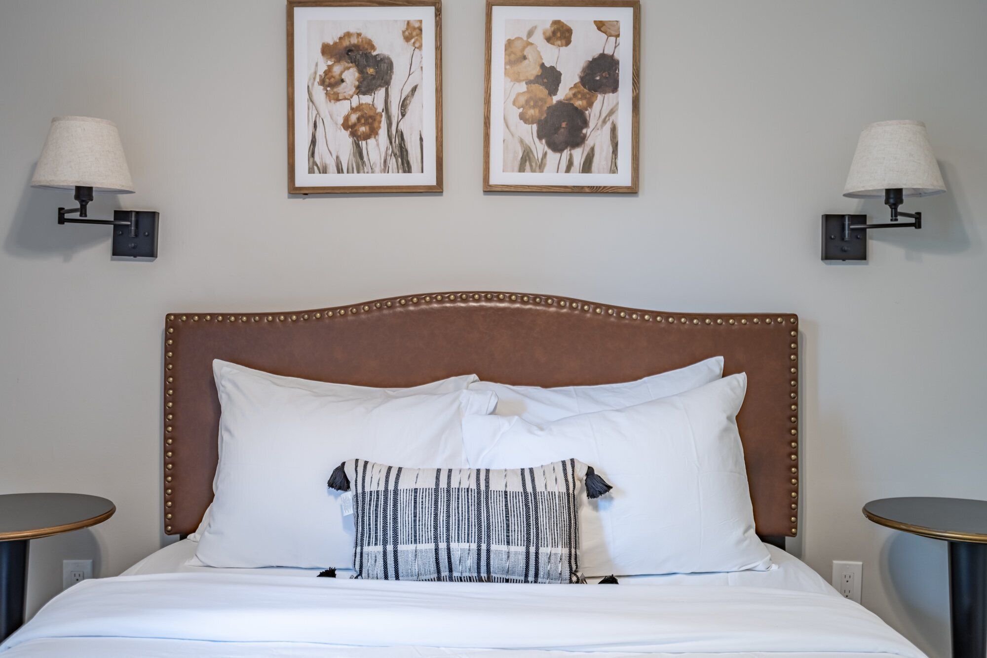 A neatly made bed with a brown upholstered headboard, white pillows, and a patterned throw. Two framed floral paintings hang on the wall above the bed.