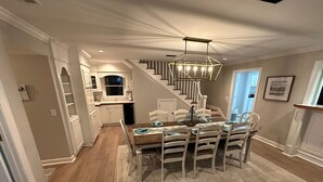 A dining area with a wooden table set for six, surrounded by white chairs, a chandelier above, and a staircase leading to an upper level.