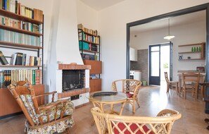 Een woonkamer met een open haard, boekenkasten, rieten meubels en een zichtbare keuken door een open deur.