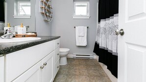 Une salle de bain avec un lavabo blanc, des toilettes et un rideau de douche. Il y a deux fenêtres et un porte-serviettes.