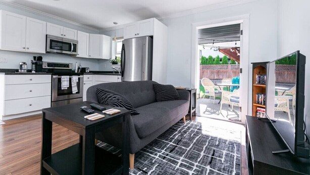 Une cuisine moderne avec des meubles blancs, un réfrigérateur en acier inoxydable et un four à micro-ondes. On y trouve un canapé de couleur foncée et une petite table sur laquelle reposent une télécommande et un livre.