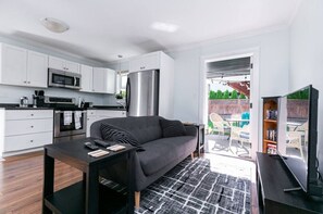 A modern kitchen with white cabinets, a stainless steel refrigerator, and a microwave. There is a dark-colored sofa and a small table with a remote control and a book.