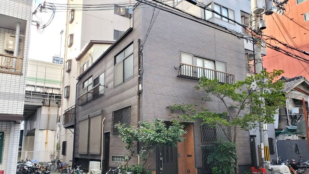 A modern building with a brick facade, a black door, and a small balcony. There are bicycles parked outside and a tree in front of the building. - Matsuyamachi Dotonbori Condo (Osaka)