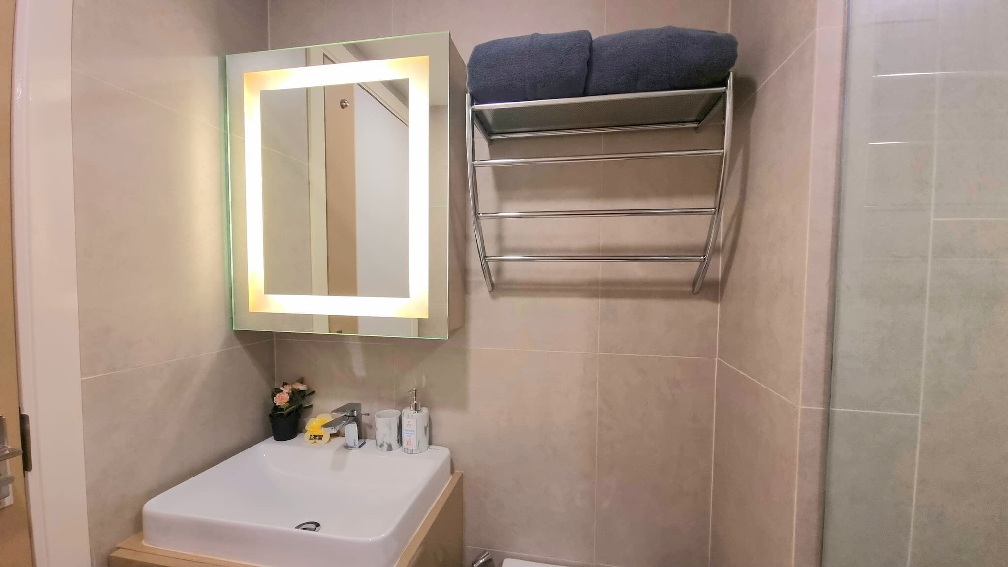 A bathroom with a white sink, a mirror with lights, a towel rack, and a small shelf with toiletries.