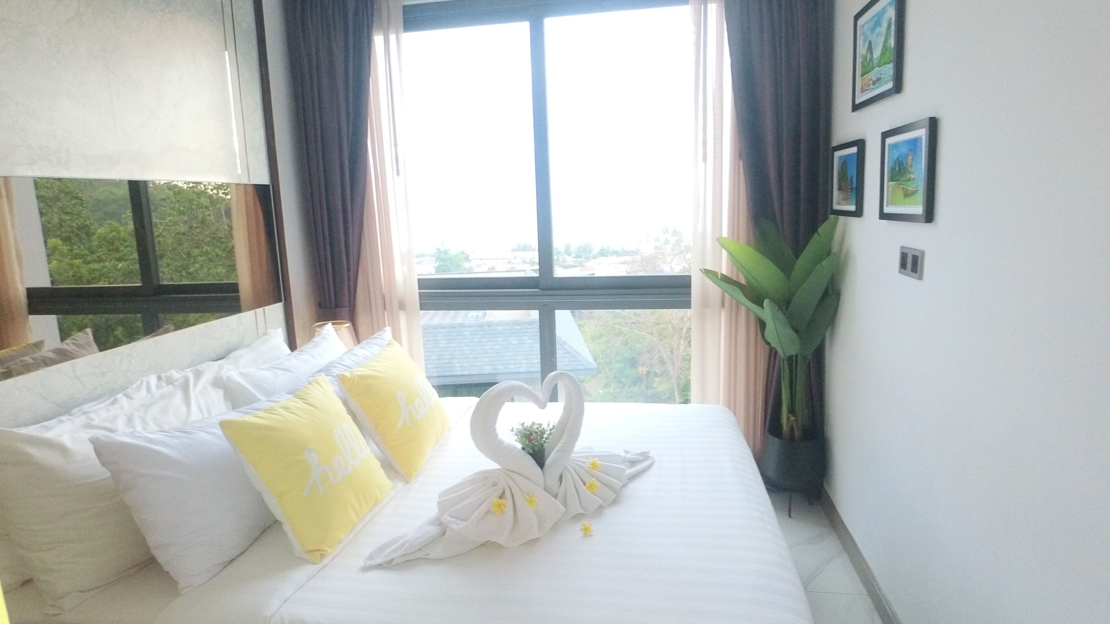 A hotel room with a bed, pillows, a heart-shaped decoration, a window, curtains, and a plant.