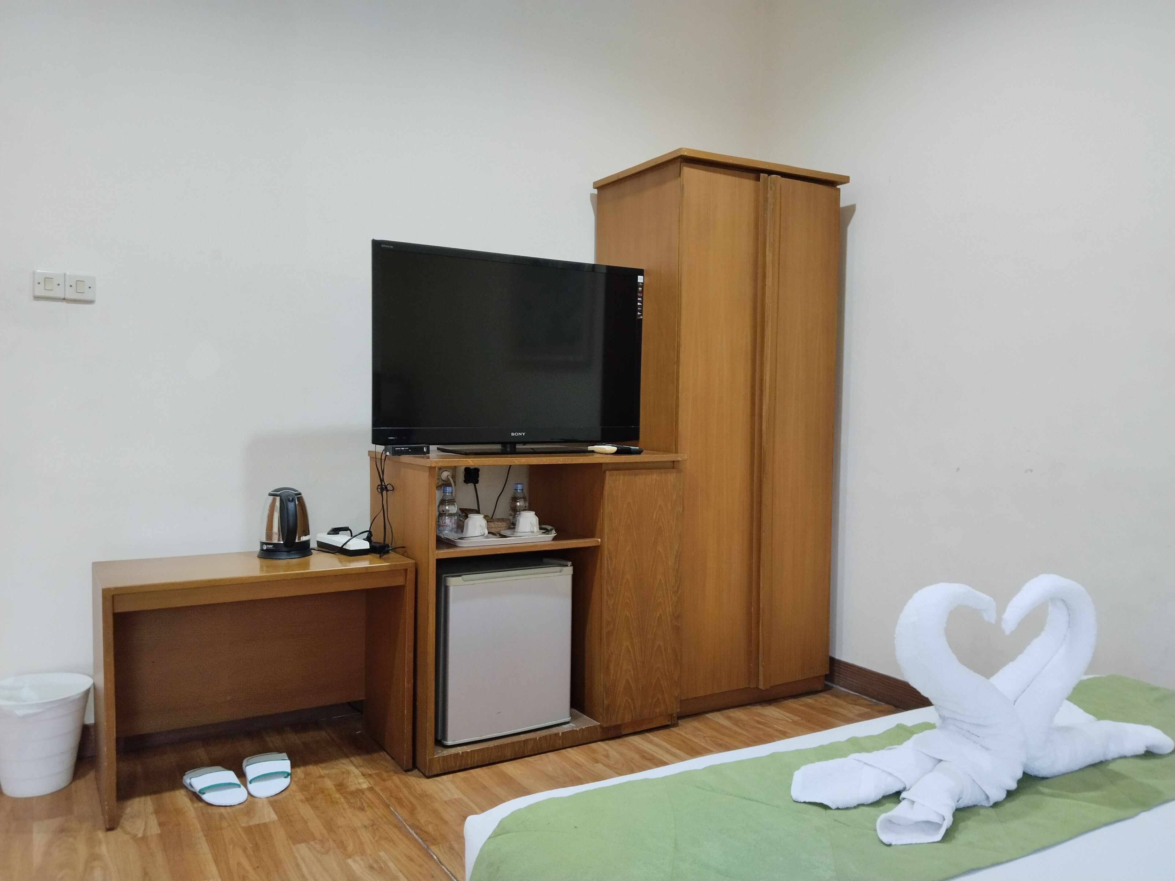 A small room with a wooden desk, a television, a mini-fridge, a cabinet, a trash bin, and a pair of slippers.