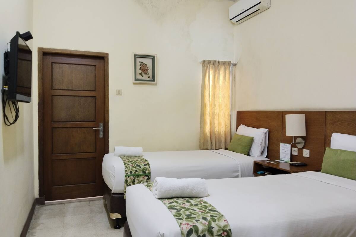 A hotel room with two single beds, a wooden door, an air conditioning unit, a small table with a lamp, and a framed picture on the wall.