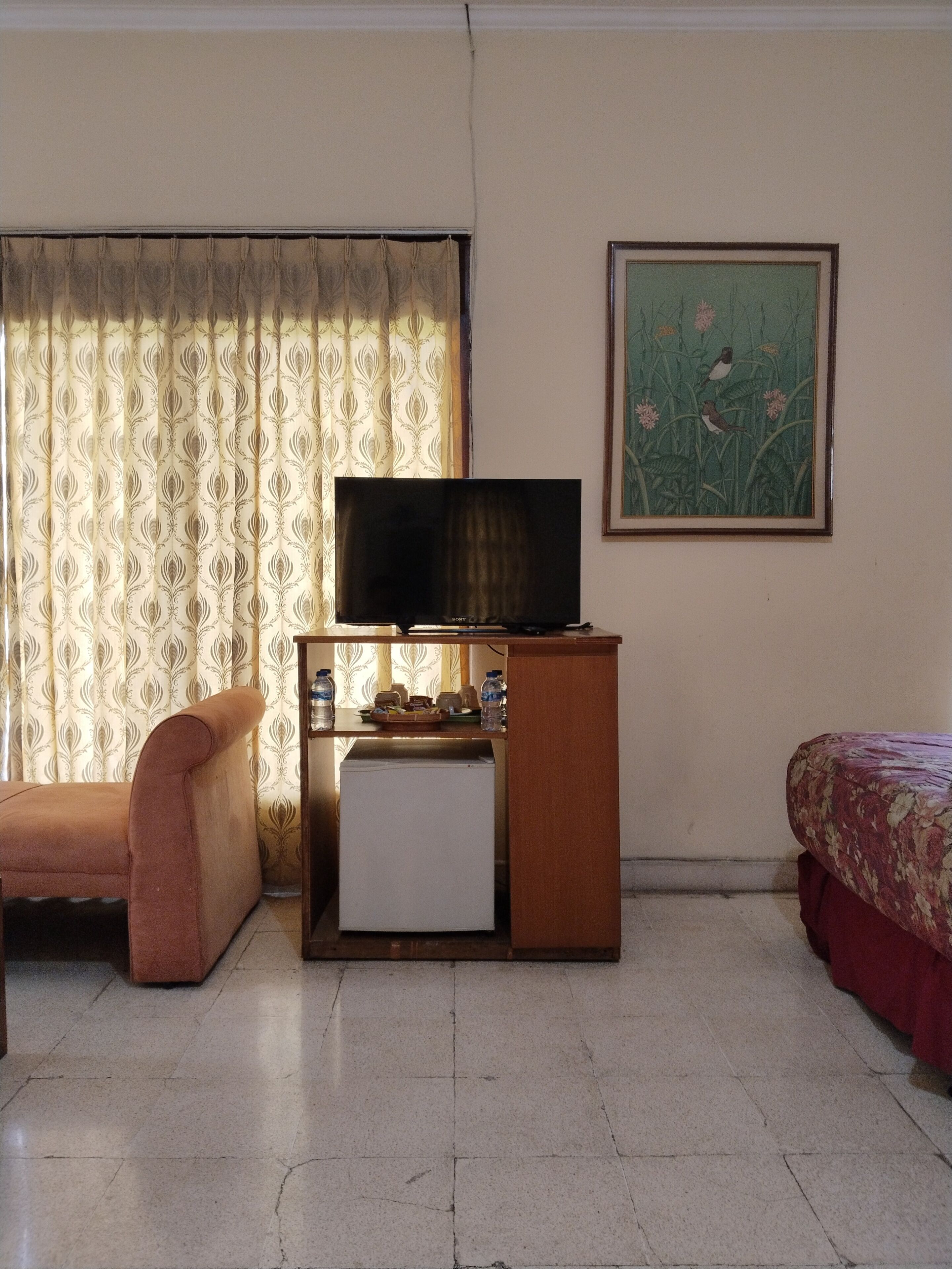 A hotel room with a bed, a sofa, a TV on a stand, a small fridge, and a painting on the wall.