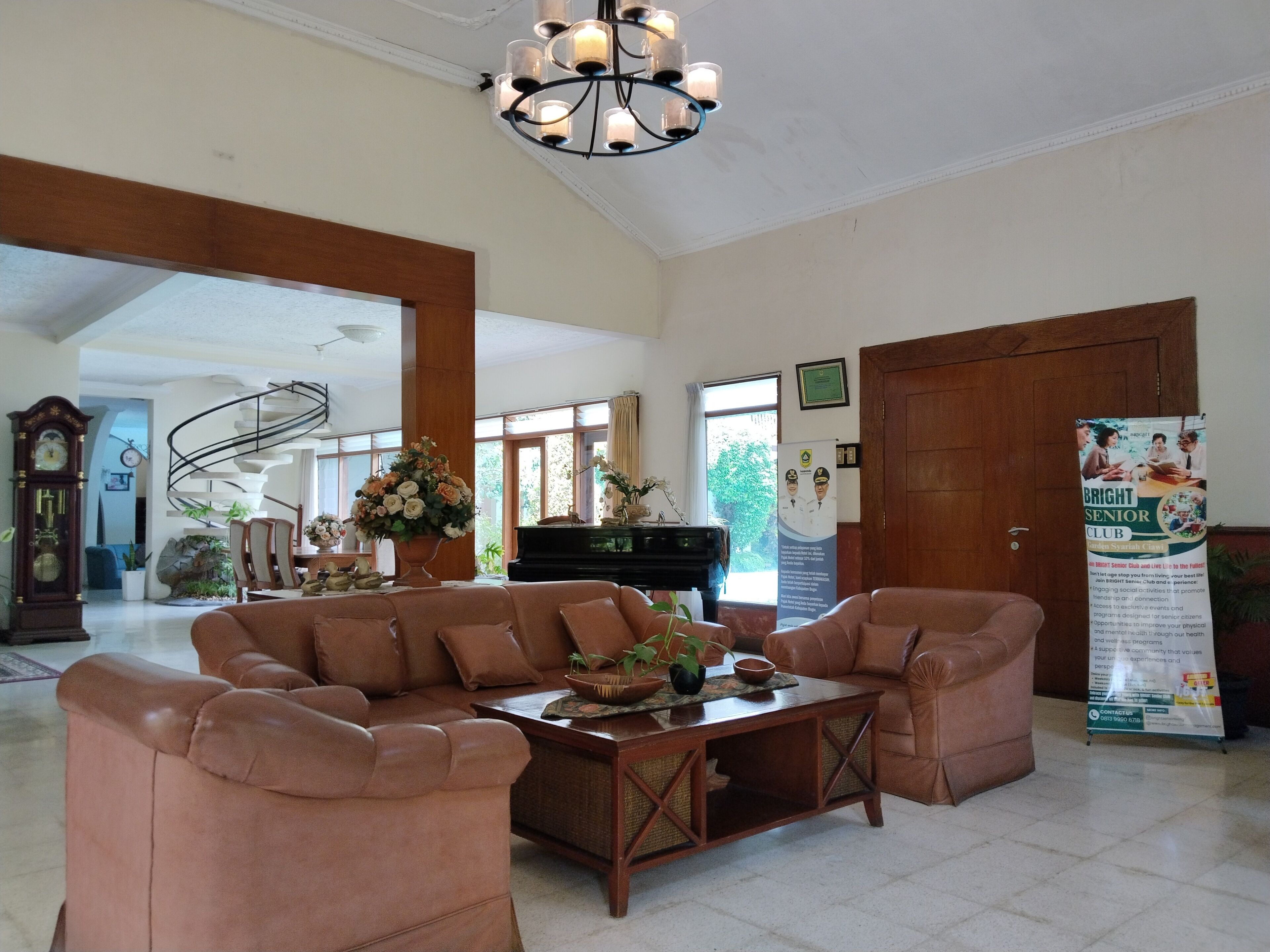 A spacious living area with a brown leather sofa, a wooden coffee table, and a chandelier. There is a piano and a large window providing natural light.
