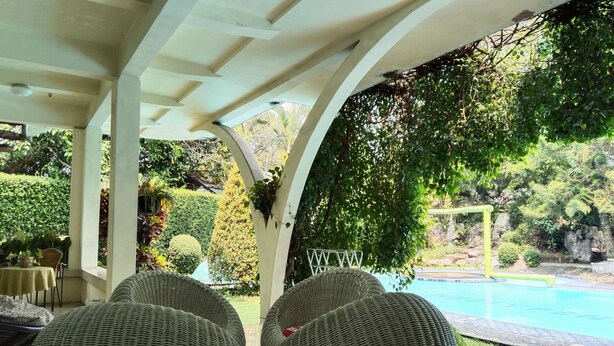A covered outdoor seating area with wicker furniture, a pool, and lush greenery. - The Garden Family Guest House powered by Cocotel. (Ciawi)