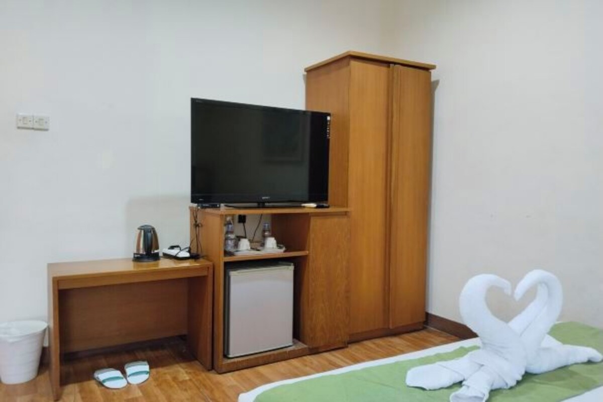A small room with a wooden desk, a television, a small refrigerator, and a bed with a green bedspread.