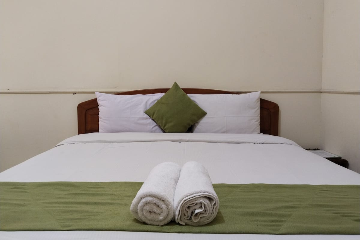 A neatly made bed with white bedding, a green throw blanket, and two rolled white towels.