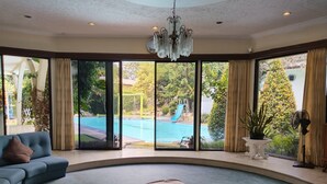 A spacious living room with a large window overlooking a swimming pool and slide, a blue sofa, and a chandelier. - The Garden Family Guest House powered by Cocotel. (Ciawi)