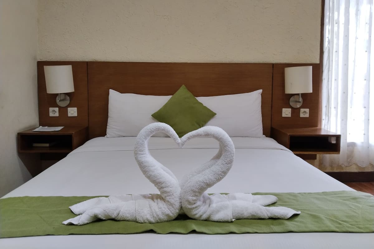 A hotel room with a neatly made bed, a green throw blanket, and two white towels arranged in the shape of swans on the bed.