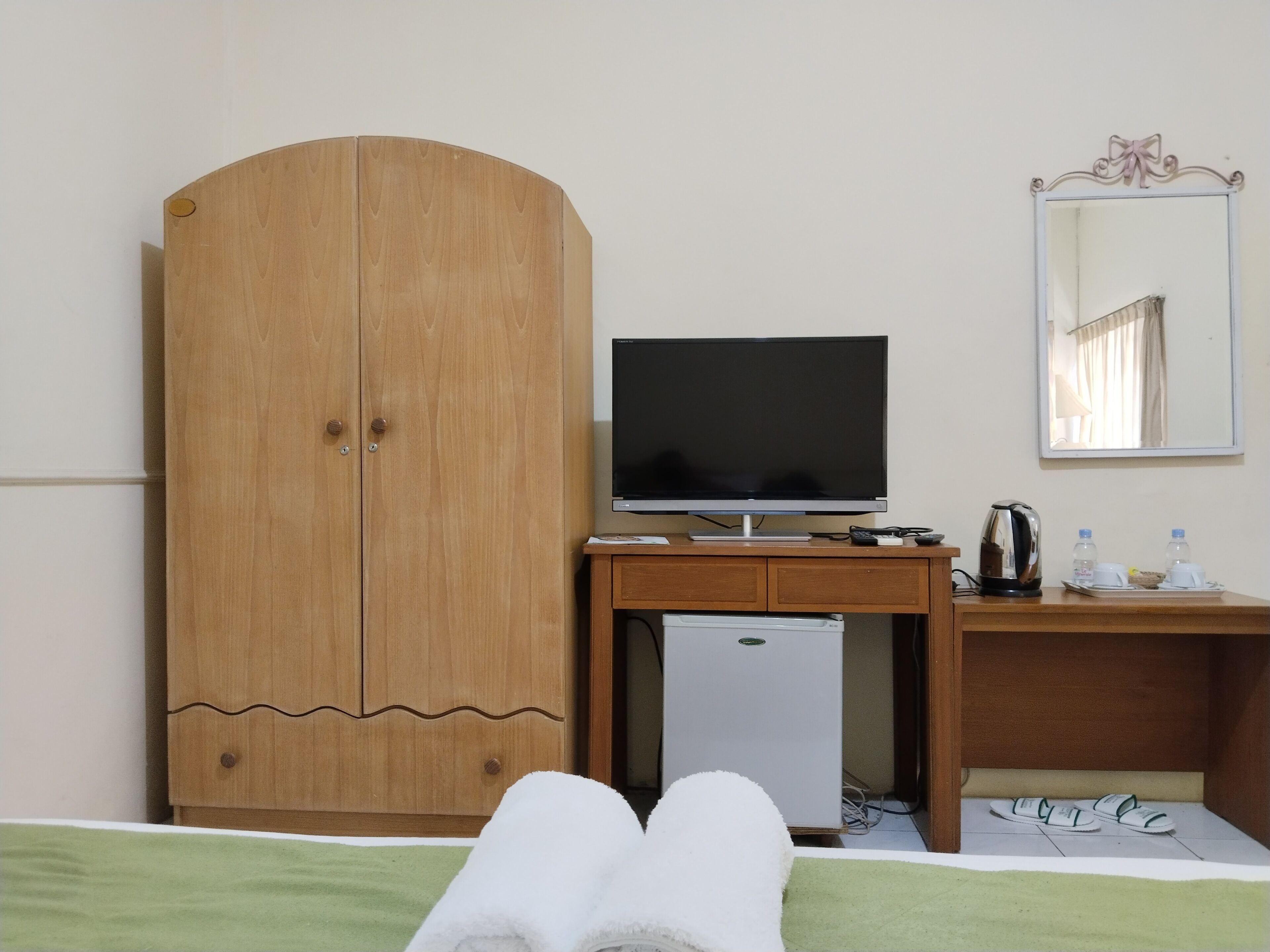 A hotel room with a bed, a wooden wardrobe, a desk with a television, a mini-fridge, a mirror, and a tray with water bottles and cups.