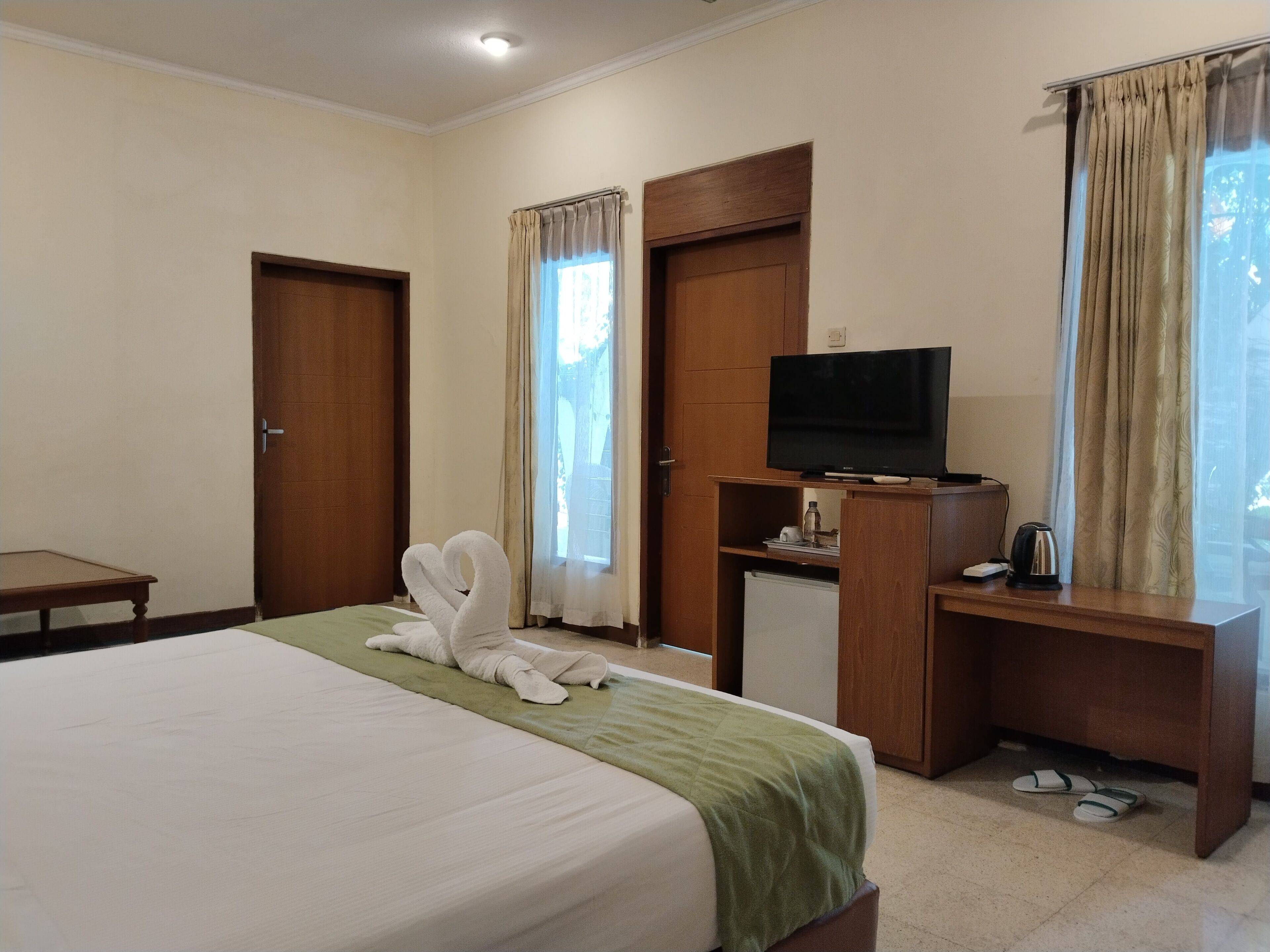 Una habitación de hotel con una cama, un televisor sobre un soporte de madera, un escritorio con un hervidor y un par de zapatillas en el suelo.