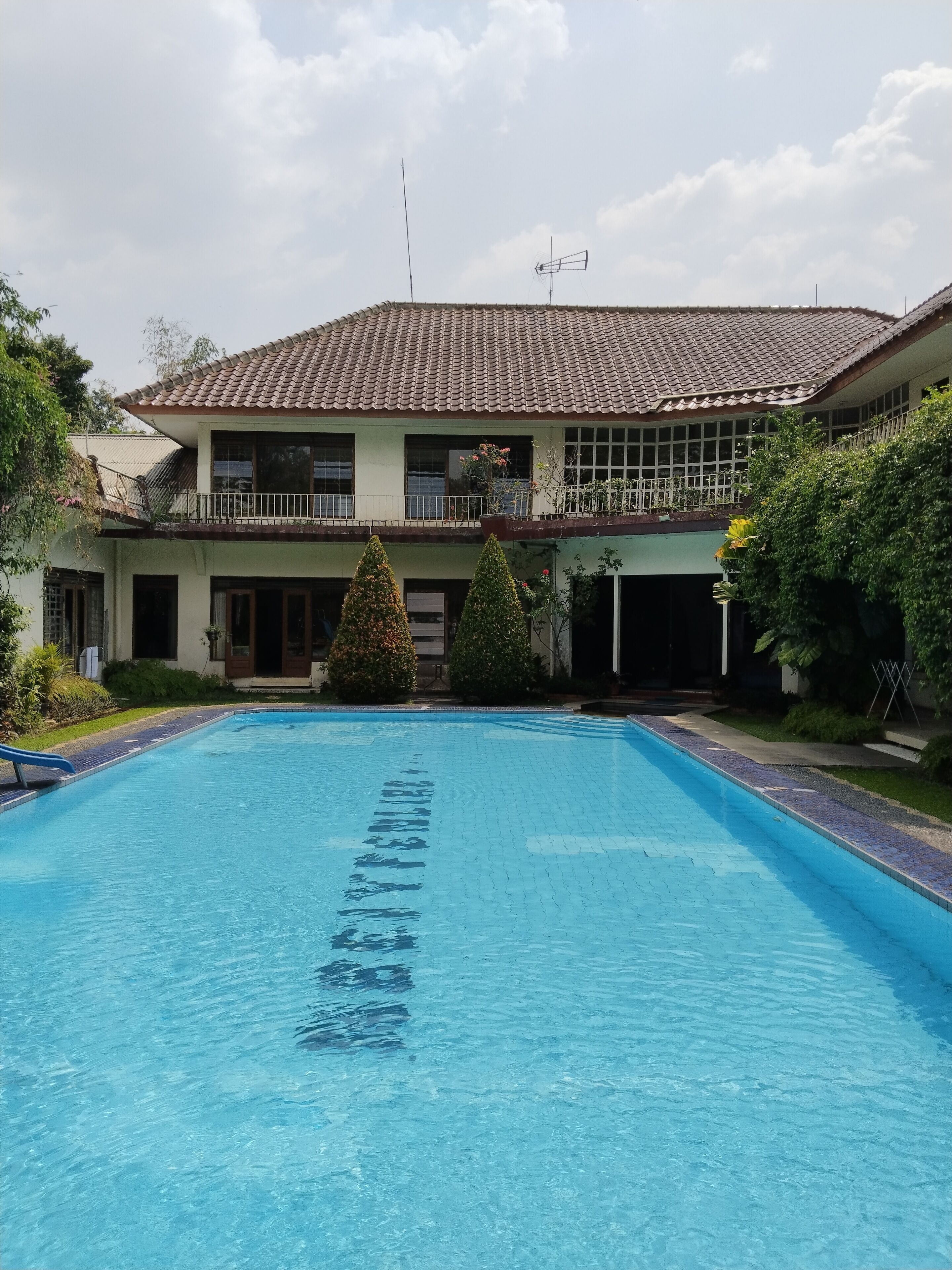 Une piscine dotée d’une terrasse carrelée, entourée d’un bâtiment à deux étages, lui-même surmonté d’un toit et d’un balcon carrelés.