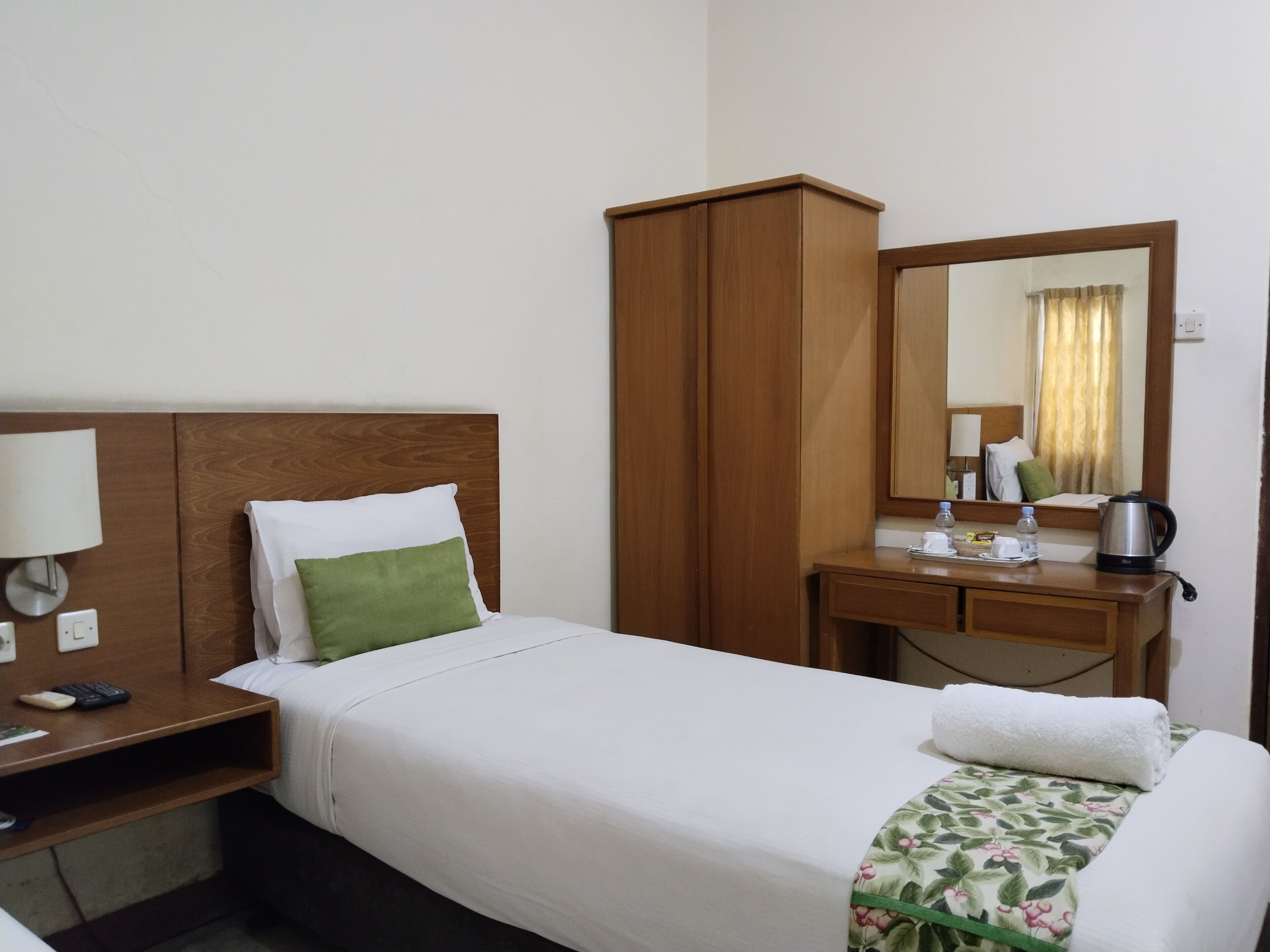 A hotel room with a bed, a wooden wardrobe, a bedside table with a lamp, a mirror, and a small desk with a kettle and a bottle.