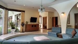 A spacious living room with a curved seating area, a wooden cabinet, a flat-screen TV, and a chandelier. - The Garden Family Guest House powered by Cocotel. (Ciawi)