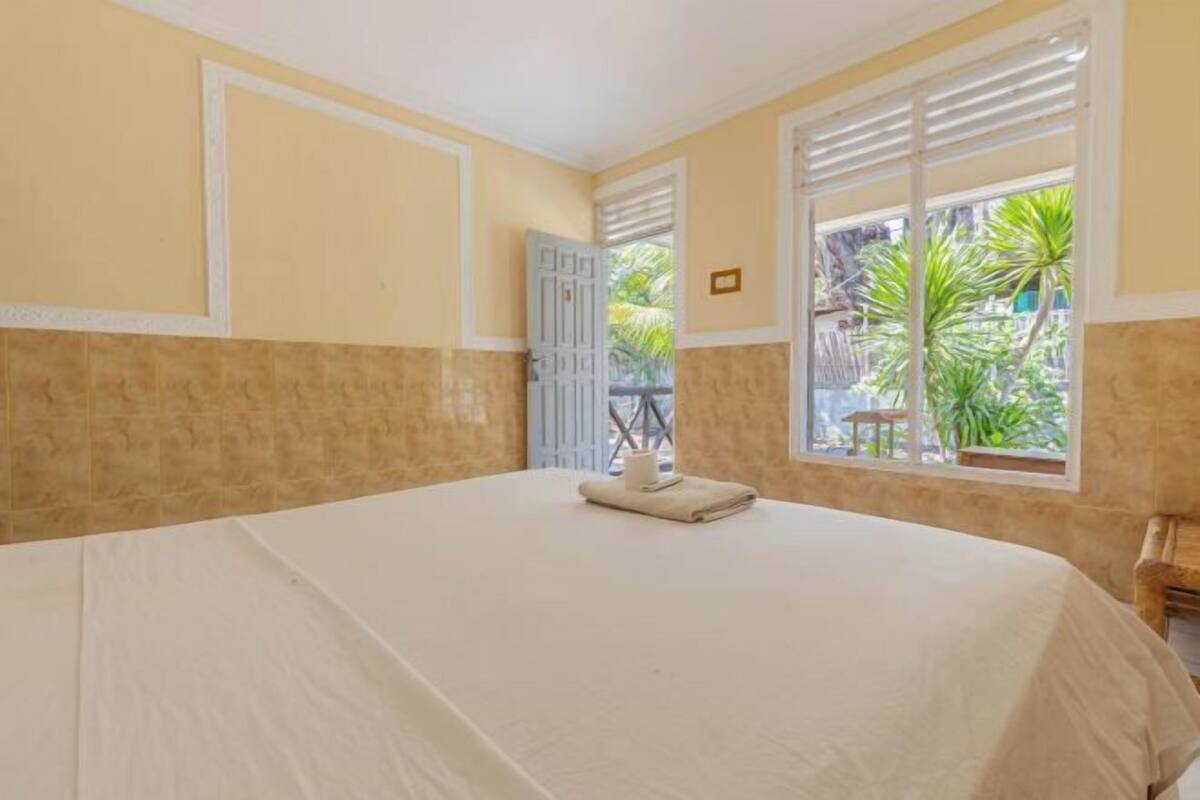 A room with a bed, a window with blinds, a door, and a view of greenery outside.