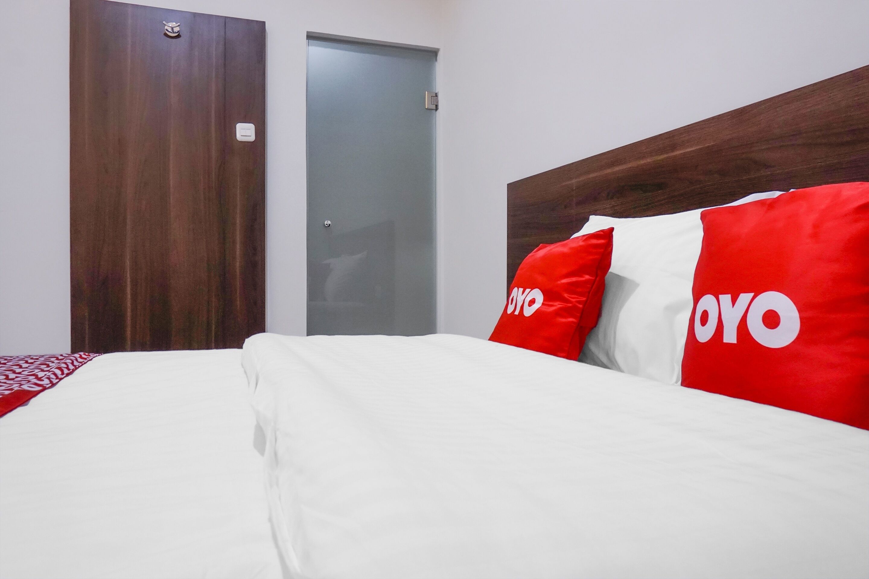 A hotel room with a wooden headboard, a bed with white bedding, and red and white pillows with the word "OYO" on them.