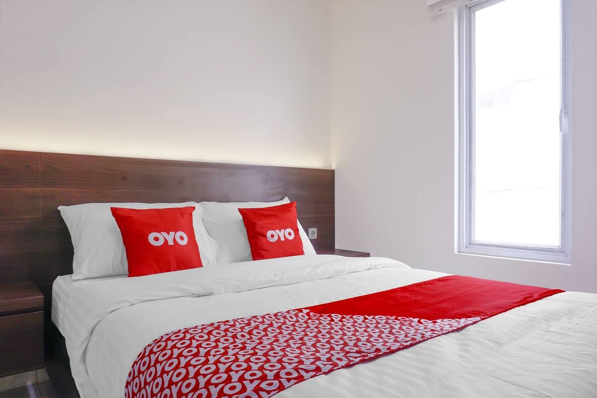A hotel room with a bed, two red pillows with the word "OYO" on them, a white bedspread, and a window with daylight coming through.