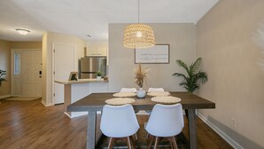 A dining area with a wooden table, white chairs, a woven pendant light, and a framed quote on the wall.