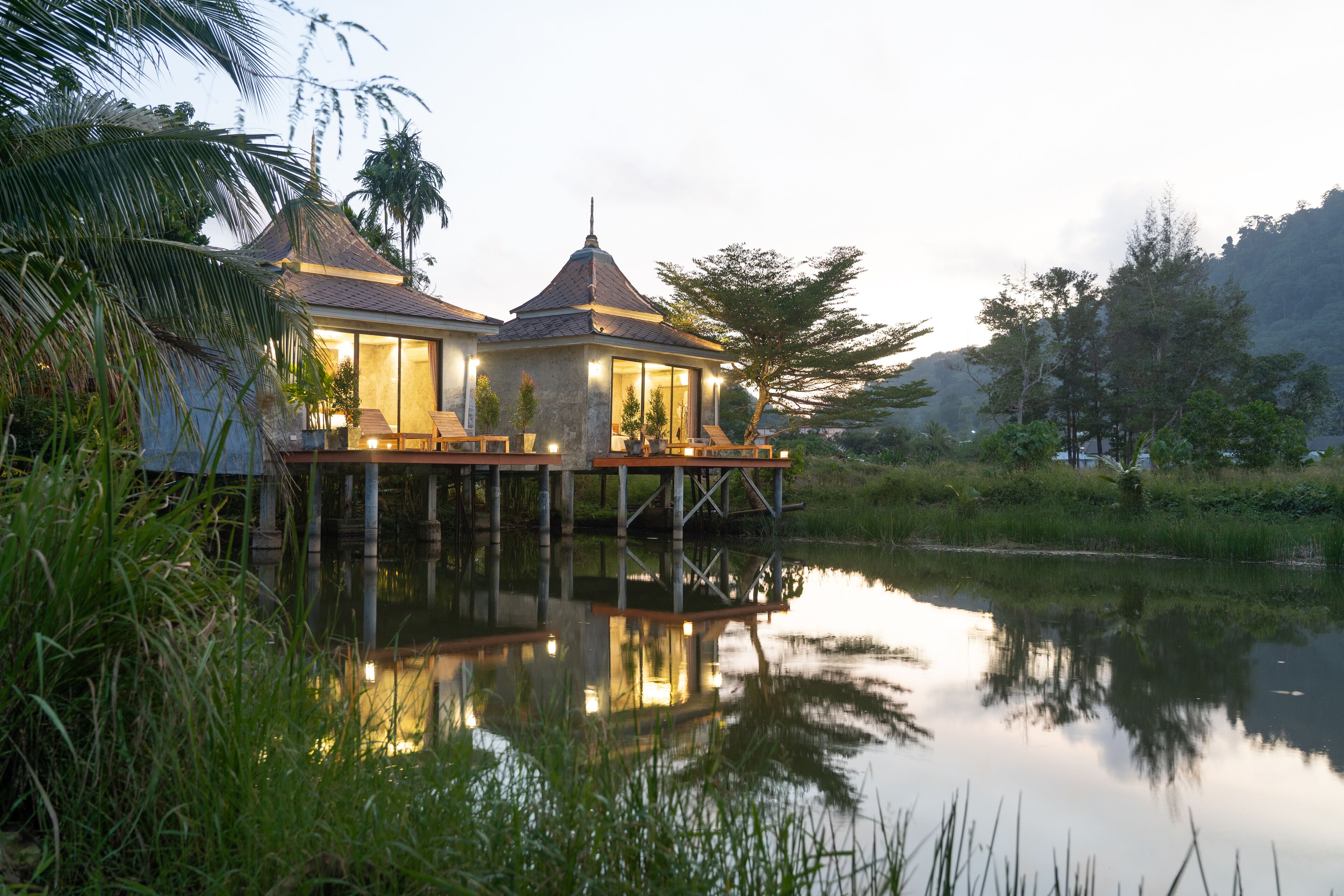 A house with a thatched roof, elevated on stilts over water, with a wooden deck and a small pavilion.