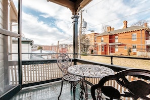 A covered balcony with a wrought iron table and chairs, overlooking a brick building and a white picket fence.