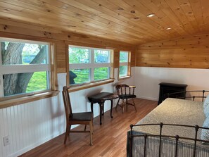 Una habitación con techo y paredes de madera, ventanas amplias, una cama, una mesa y dos sillas.