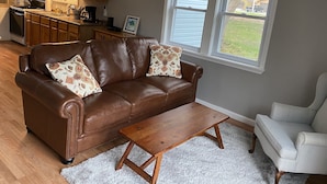 Un salon avec un canapé en cuir marron, une table basse en bois et un fauteuil blanc. On aperçoit une cuisine à l’arrière-plan, avec des meubles en bois et une fenêtre.
