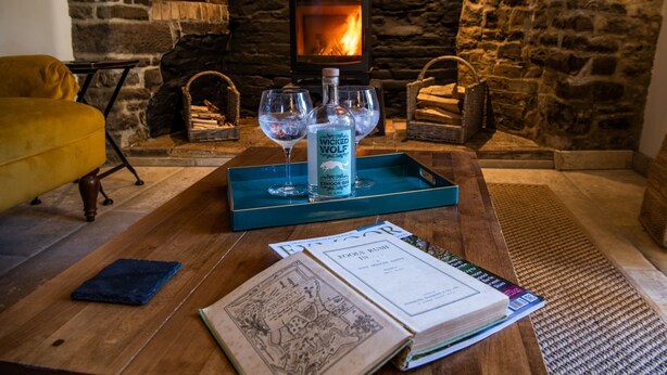 A cozy room with a stone fireplace, a wooden table with a glass bottle, wine glasses, and books. - Keepers Lodge Knowstone (Knowstone)