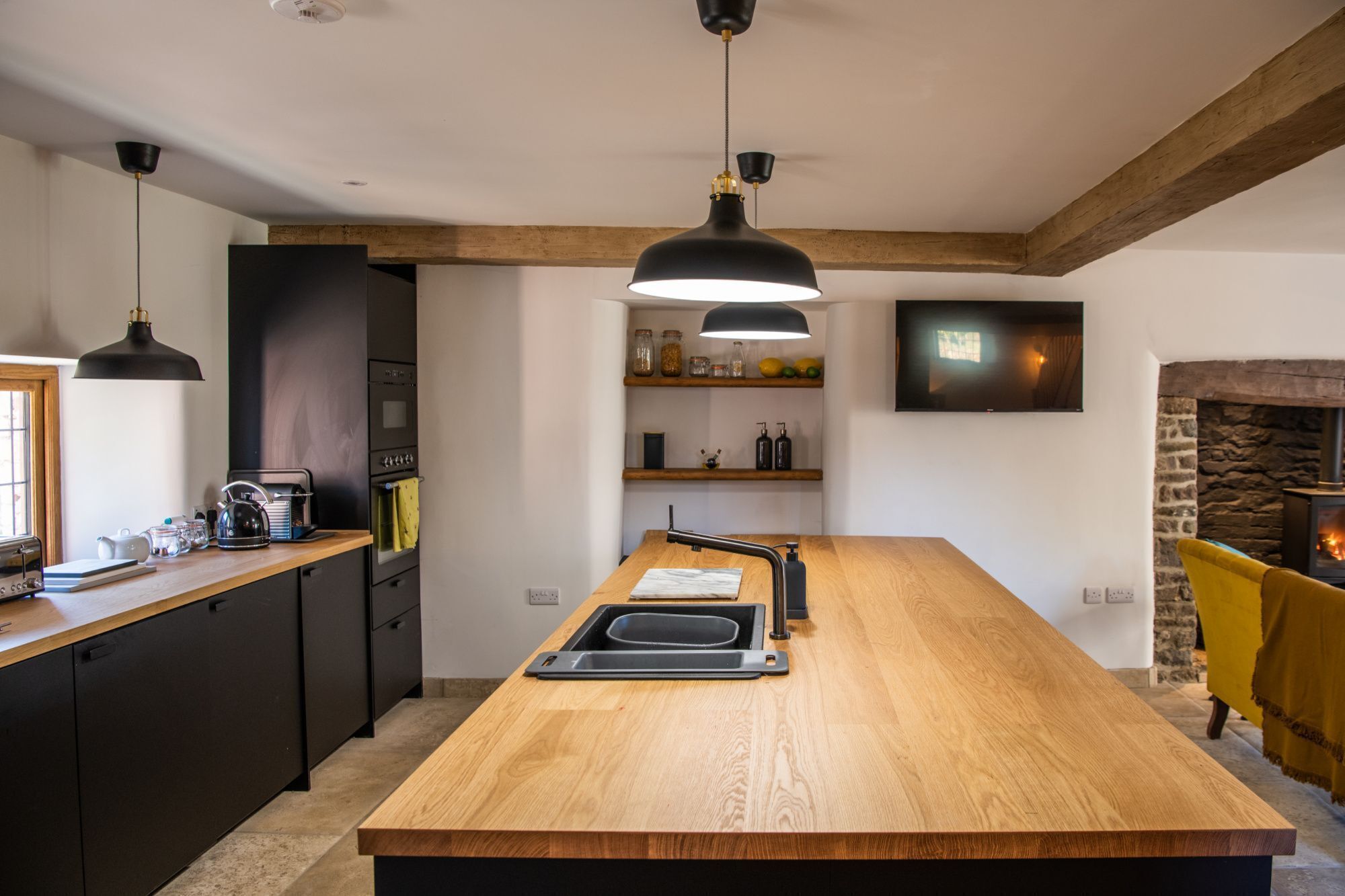 A modern kitchen with a central island, black pendant lights, a stone fireplace, and a TV mounted on the wall.