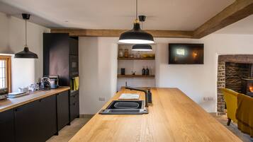 A modern kitchen with a central island, black pendant lights, a stone fireplace, and a TV mounted on the wall.