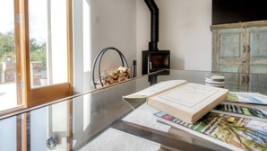 Una stanza con un tavolo in vetro su cui sono esposti un libro e riviste, un camino a legna nero e un mobile rustico.