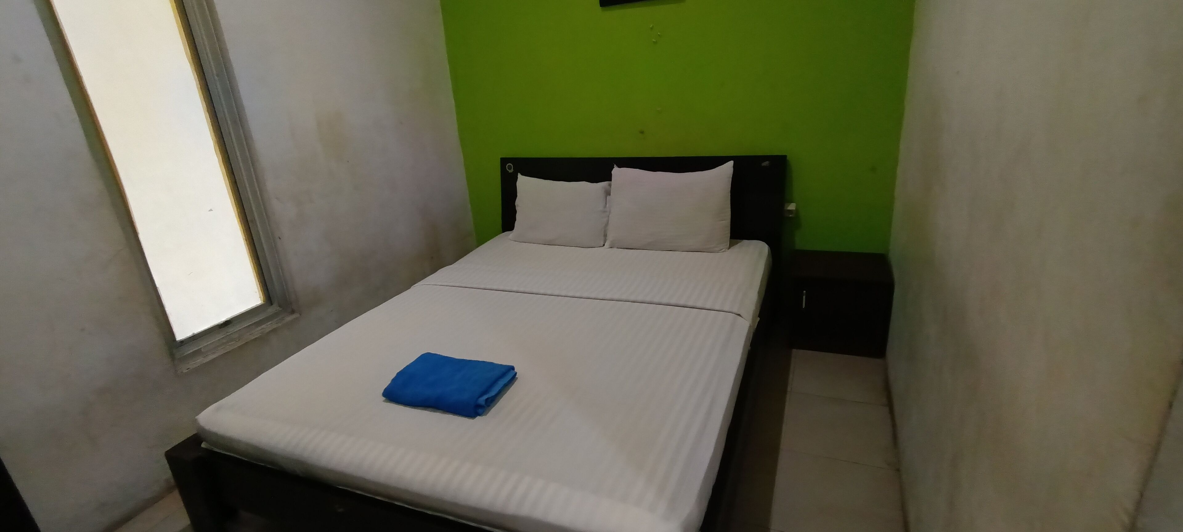A small, single-bed room with a green wall, a window, and a neatly made bed with a blue towel.