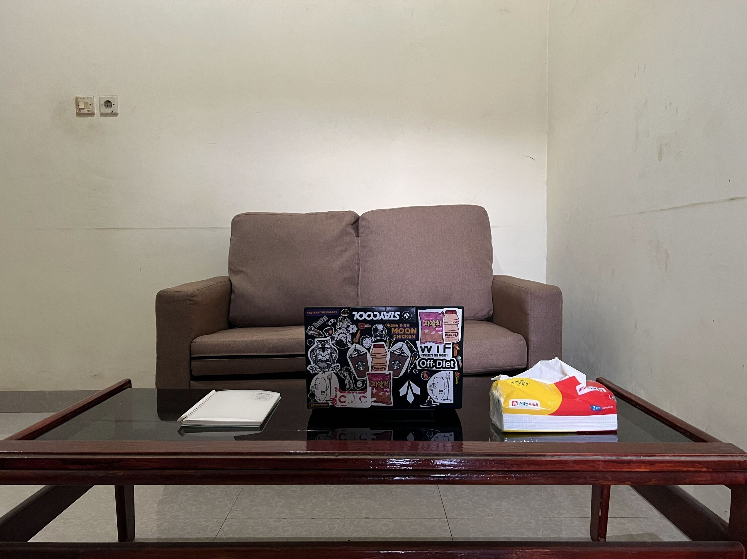 Un sofá marrón frente a una pared lisa, una mesa de centro de vidrio con una libreta, un tablero cubierto de pegatinas y un paquete de bocadillos.
