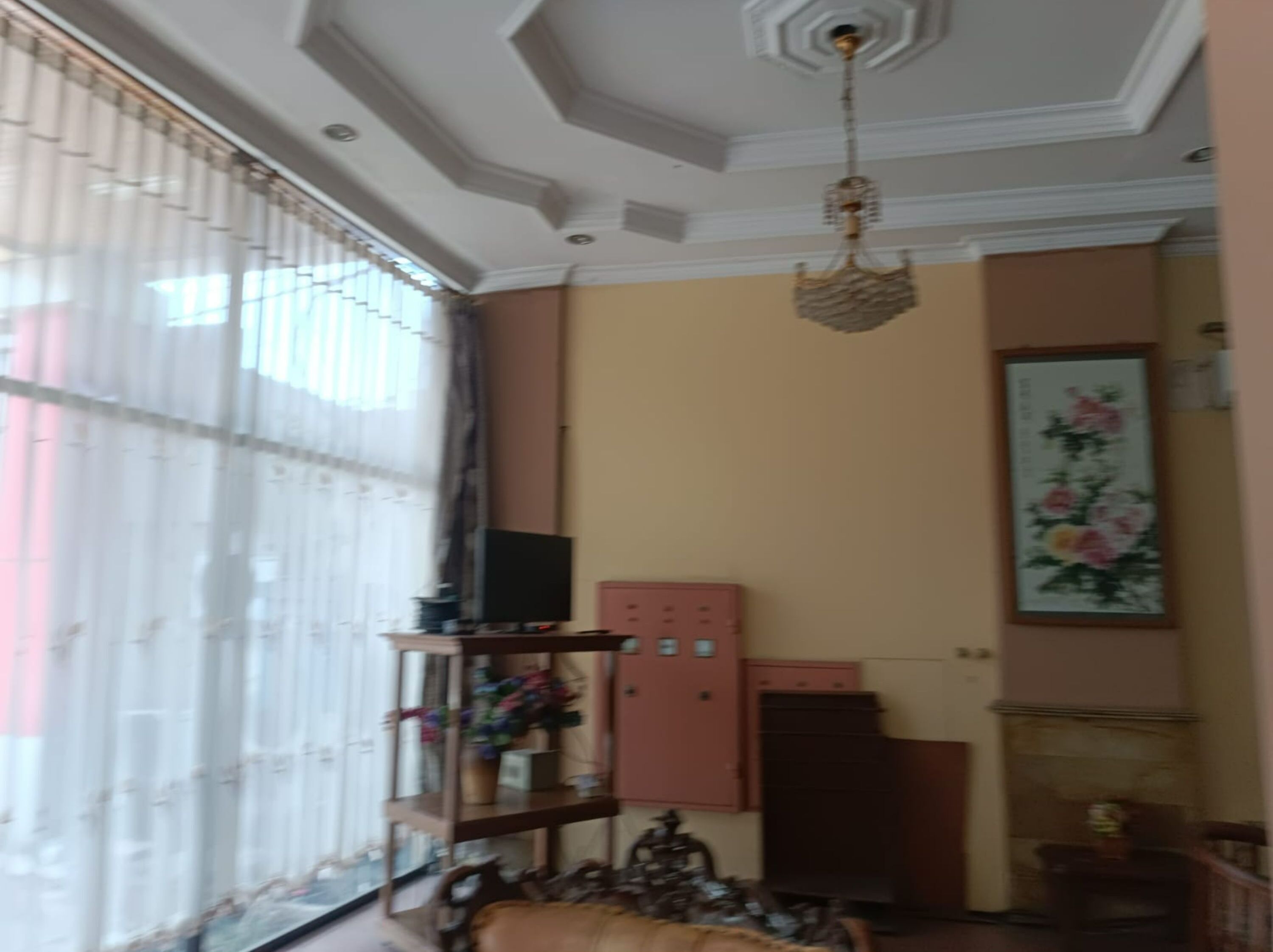 Una habitación con un ventanal grande, una araña, un televisor sobre un mueble, un armario y un cuadro en la pared.