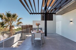 A modern outdoor patio with a dining table and chairs, a pergola, and a view of palm trees and the sea.